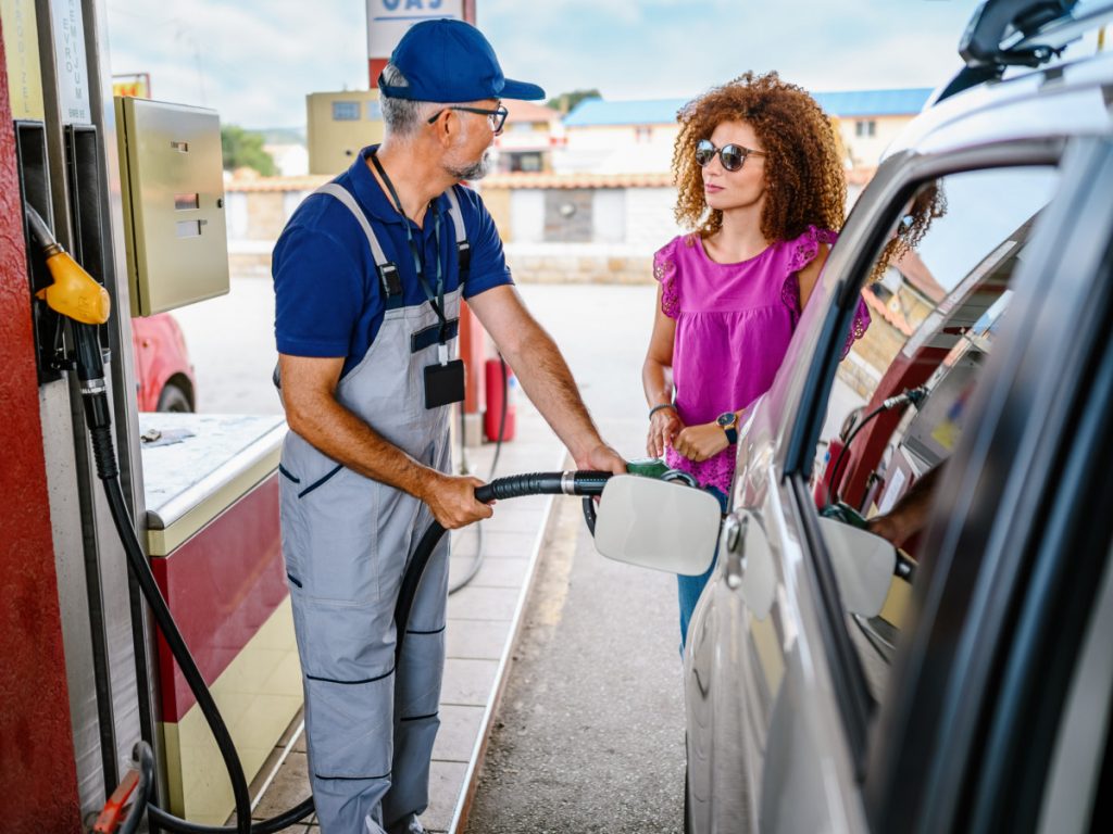 A partir de hoje, 1° de fevereiro de 2025, os preços dos combustíveis estão mais caros. Na foto, o frentista, de macacão claro, de camisa e boné azul abastecendo carro de motorista mulher que está com camisa lilás e de óculos escuros.