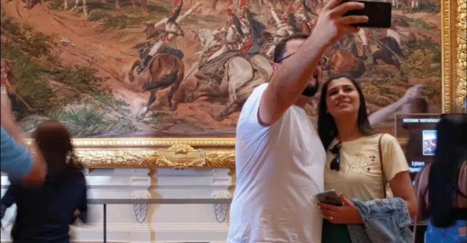 Um homem que segura o celular com uma mão, abraça sua mulher  e posam para uma selfie com o quadro, ao fundo, "Independência ou Morte", de Pedro Américo, no Museu do Ipiranga.